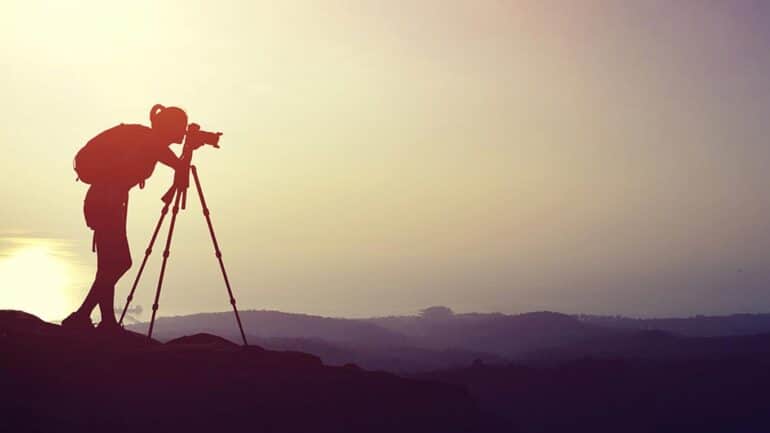 Woman photographer wearing a backpack using a tripod to take a picture of from the top of a hill